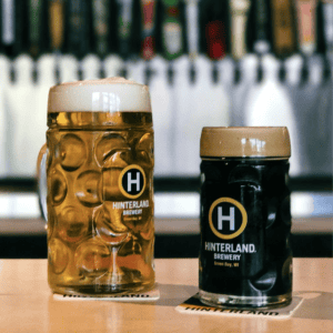 Two different sizes of Hinterland-branded glass steins full of beer sit on a bar.